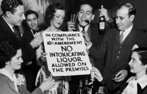A black and white photo of people gathered with wine and cigars. They hold a sign that says 'in compliance with the 18th amendment no intoxicating liquor allowed on the premises.