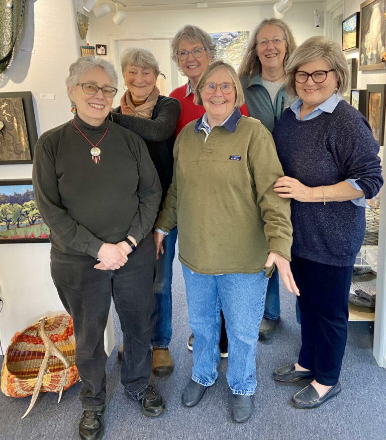 Six women smile and pose in an art gallery with art on the walls behind them.