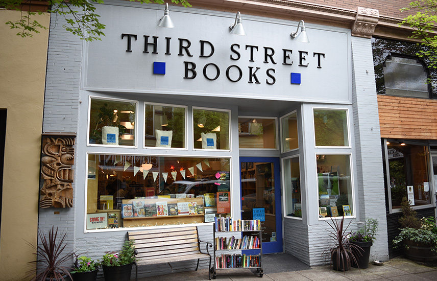 The exterior of Third Street Books.  There is a bench out front along with planters and a rack of paperback books.