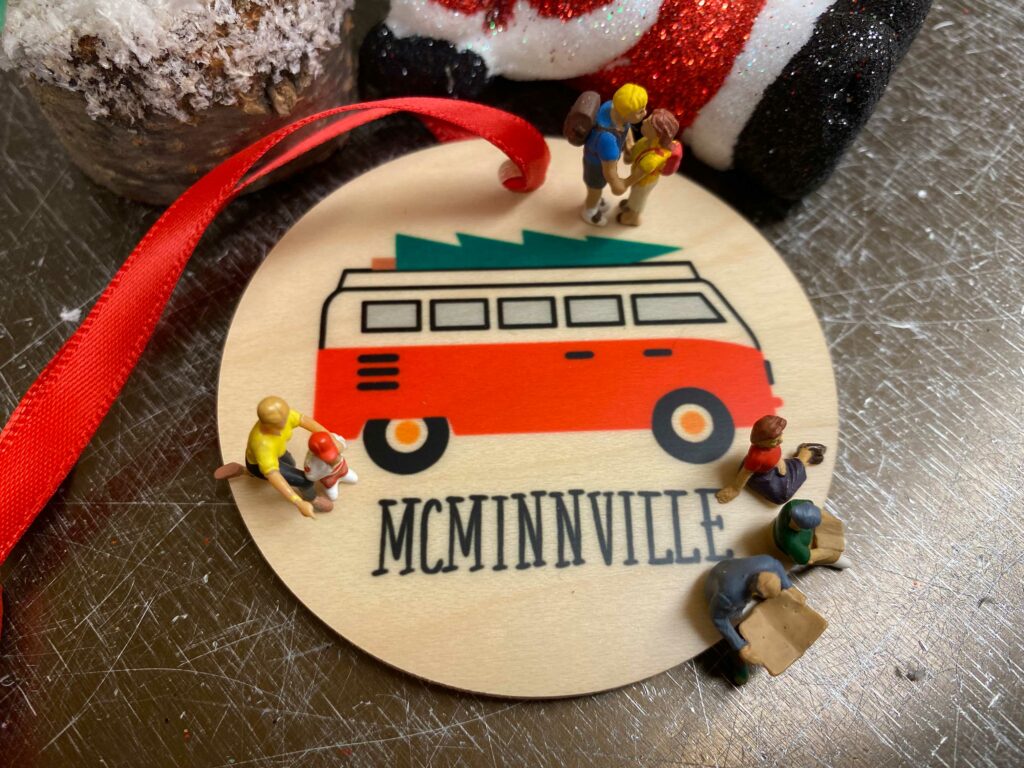The Tiny Travelers sit atop a flat wooden ornament that says McMinnville on it.