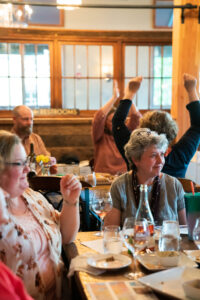 People at a table in the foreground smile. They have a bottle of pink wine, along with glasses, plates of snacks, and papers on the table. A table in the background have people that have their arms up in celebration.