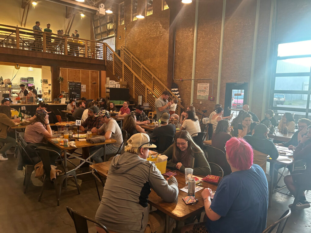 Many tables are full of people eating and drinking.  You can see that the mood is jovial.  They are in a large building with a glass garage door in the background that allows light to stream in.