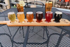 A colorful beer flight is staged on an outdoor table with two glasses of iced tea with lemon.