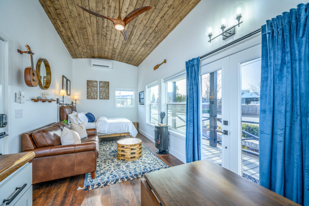 The inside of a tiny house shows white walls and a wood plank ceiling.  Blue curtains are open across french doors looking out onto a patio.