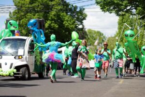 Parade participants wearing green alien suits dance in the street.