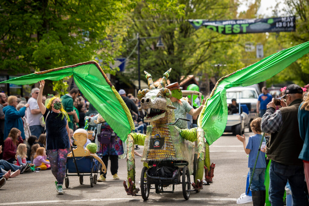 An alien sits atop a dragon contraptions, pedaling down the street during a parade.