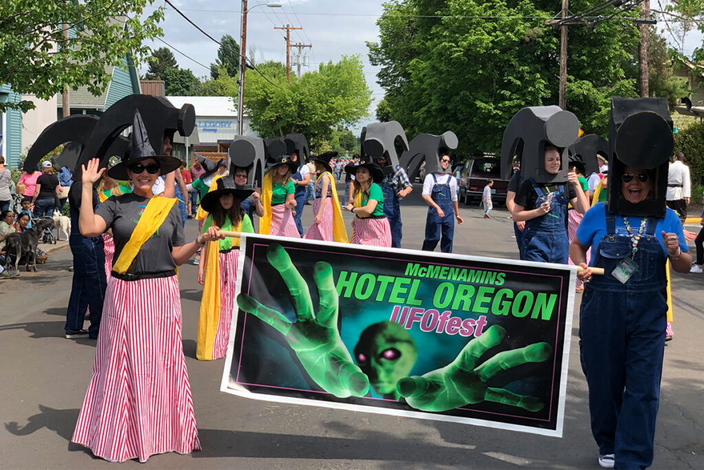 Parade participants dressed in McMenamins hammerhead and witch costumes holding a banner that says McMenamins Hotel Oregon UFOfest.