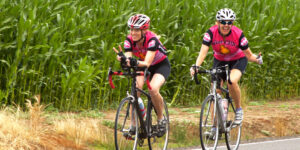 two women riding bikes on a country road in front of a corn field