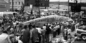 An old black and white photo of a firefighter aiming a water hose and people standing around watching.