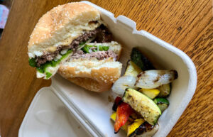 A compostable to-go container with a cheeseburger cut in half and grilled fresh vegetables.