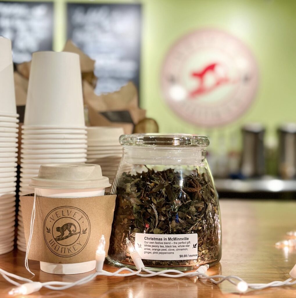 A glass jar with dried tea leaves and pink peppercorns.  It says Christmas in McMinnville.  A paper cup is next to it with a sleeve that says Velvet Monkey.  A string of white lights is place in front of them.