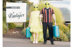 A couple dressed as aliens stand holding hands and vintage suitcases. A sign that says The Vintages is next to them.