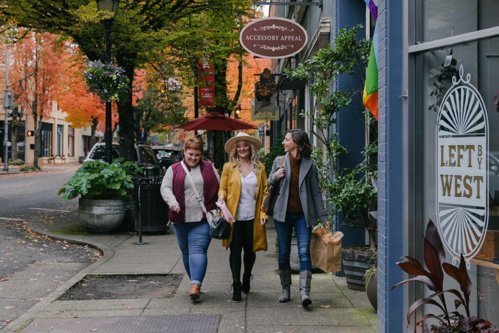 Three women walk down a quaint downtown sidewalk, smiling and chatting with shopping bags in their hands.