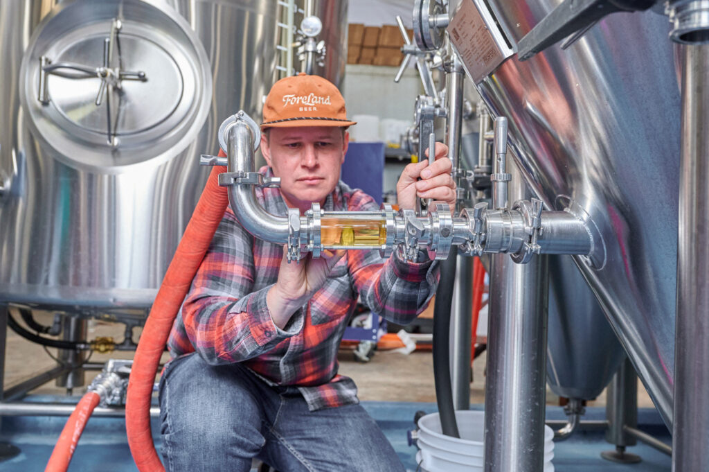 A person with a ForeLand Beer hat on crouches down to inspect beer brewing equipment.