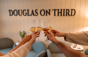 4 people cheers glasses of white wine. A sign on the wall behind them says Douglas on Third.
