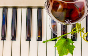 A glass of red wine rests on a piano keyboard. There is a grape vine next to it.