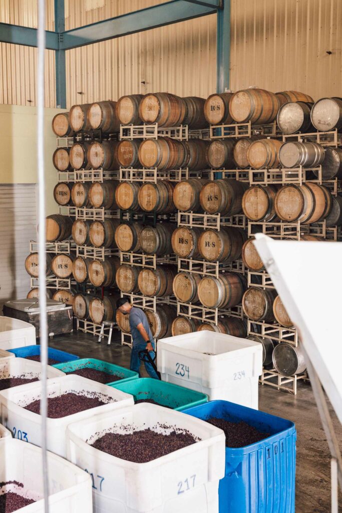 There are several large plastic containers full of what looks like grape pumice.  A wall of wine barrels is in the background.  A man wheels one of the containers away from the camera view.