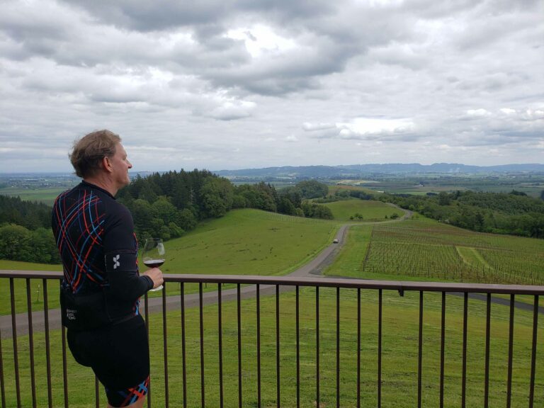 A man holds a glass of wine as he looks out at a grassy, expansive view of the Willamette Valley.