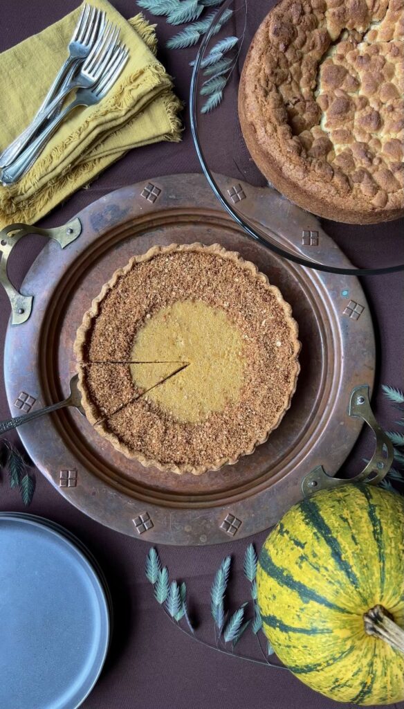 A pumpkin pie with a slice cut into it, ready to be pulled out with a pie server.  Another pie is next to it, uncut.  And forks are placed on a pile of gold napkins.