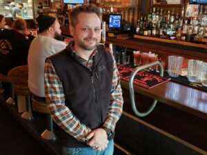 Justin Dillingham stands smiling in front of the bar. Three prepared bloody marys are in the background along with a full bar of spirits.