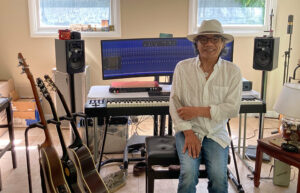 A man with a hat and glasses sits on a piano bench and smiles. His piano studio is behind him.