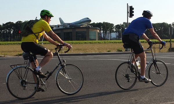 Two people wearing bicycle helmets ride their bikes down a road. A large building with a jet on top is in the background.