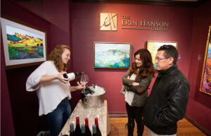 A person pours wine into glasses and they talk to a smiling couple. Art is on the walls in the background.