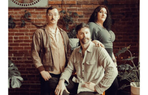 Three people in neutral colored clothing stand and pose together.