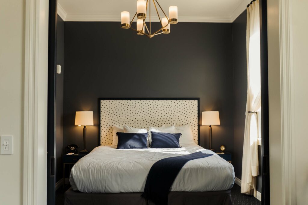 Sun shines through a tall long window onto a luxe bed.  There are side tables with lamps on either side of the bed and a pretty chandelier hanging from the ceiling.