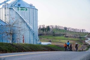 A group of cyclists ride past a grain silo with a vineyard in the background.