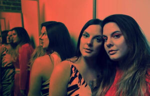 A photo of two women standing together with a mirror behind them. The photo has a red/orange tint.