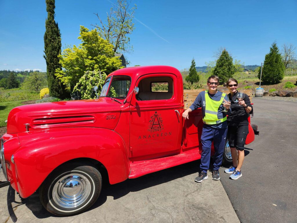Two people pose in front of an old fashioned red truck with the logo for Anacreon winery on the truck door.  The people hold glasses of wine.