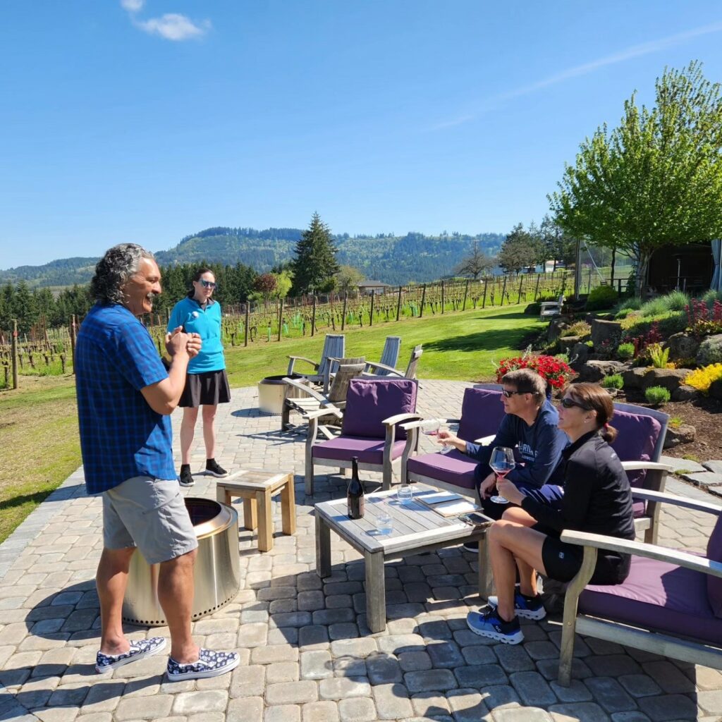Two people sit in chairs while they sip on red wine.  Two other people stand as they are talking to the seated folks.  They are outside on patio furniture with a vineyard in the background.
