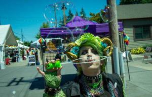 A person dressed in green costume with a green wig and golden horns blows bubbles.